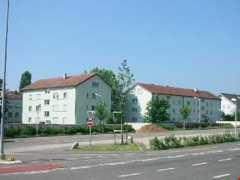Two white apartment buildings with brown roofs, streetlight, parking lot, trees, bushes, road