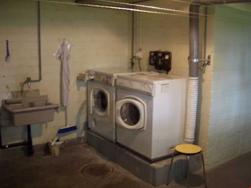 Two washing machines, sink, stool, broom, trash can, pipes