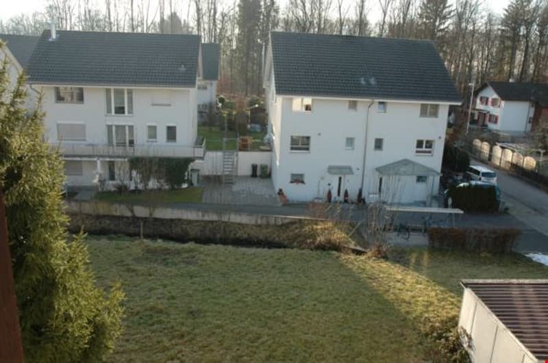 Two-story white houses with gray roofs, surrounded by trees and grass, driveway, and a small garden in front.