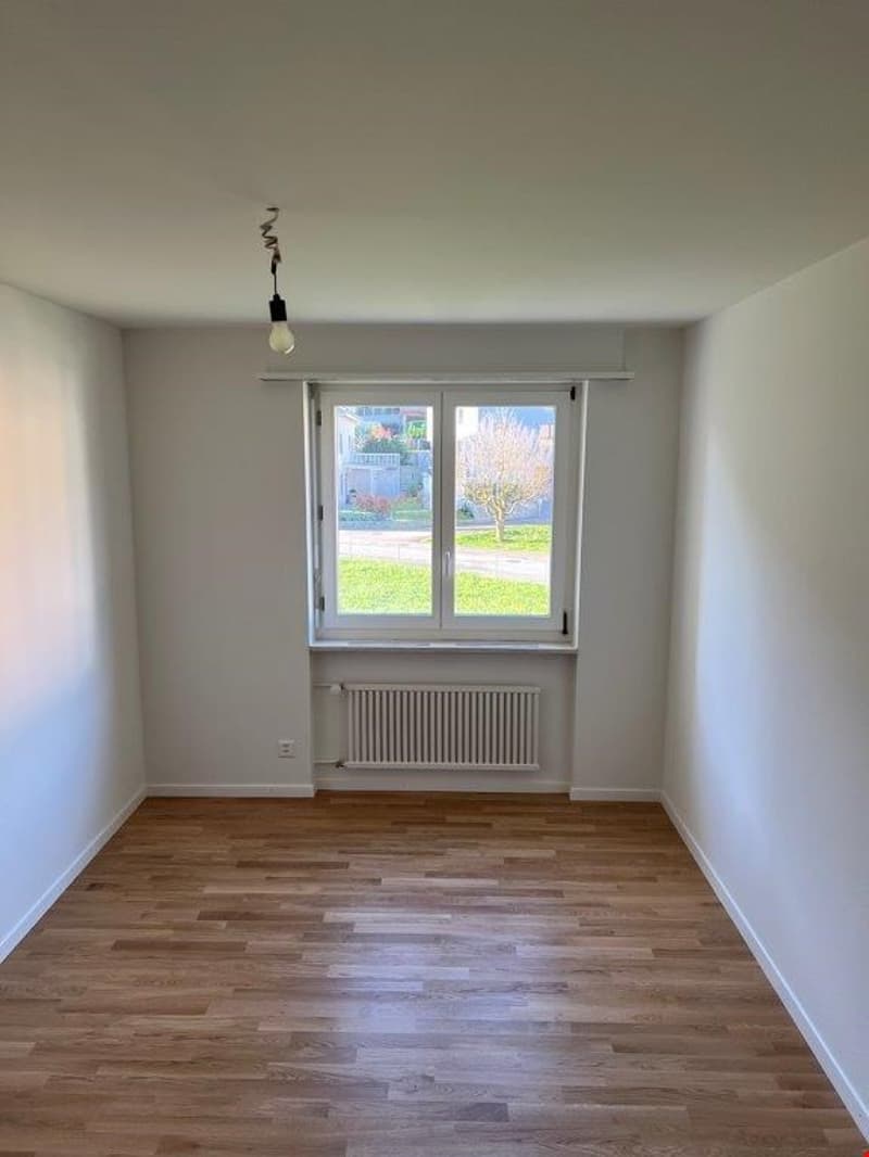 Spacious bedroom with hardwood flooring, large window overlooking a garden, and a radiator on the wall.