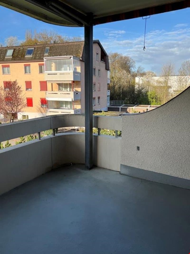 Covered balcony with concrete floor, overlooking residential buildings and trees in the background
