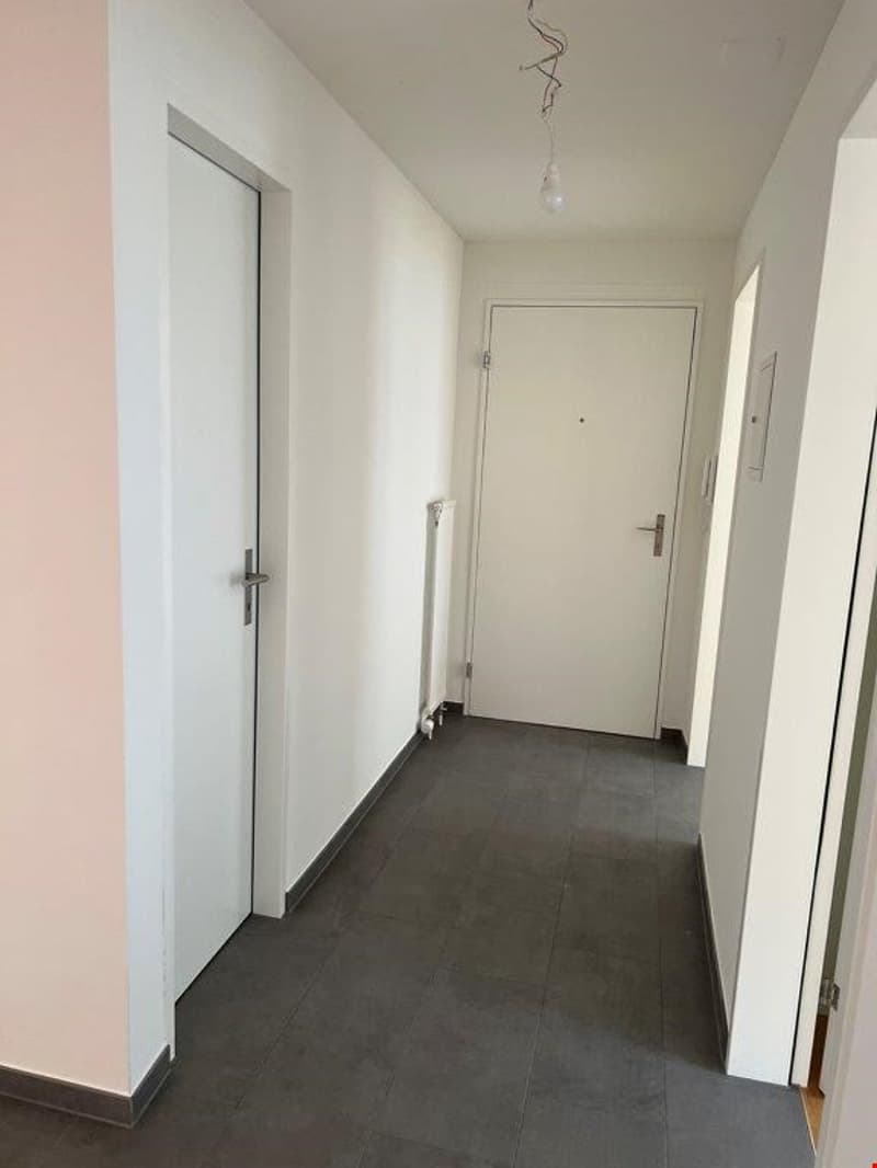 The image shows a hallway with white walls, a gray tile floor, and a white door leading to another room. There is a light fixture hanging from the ceiling.