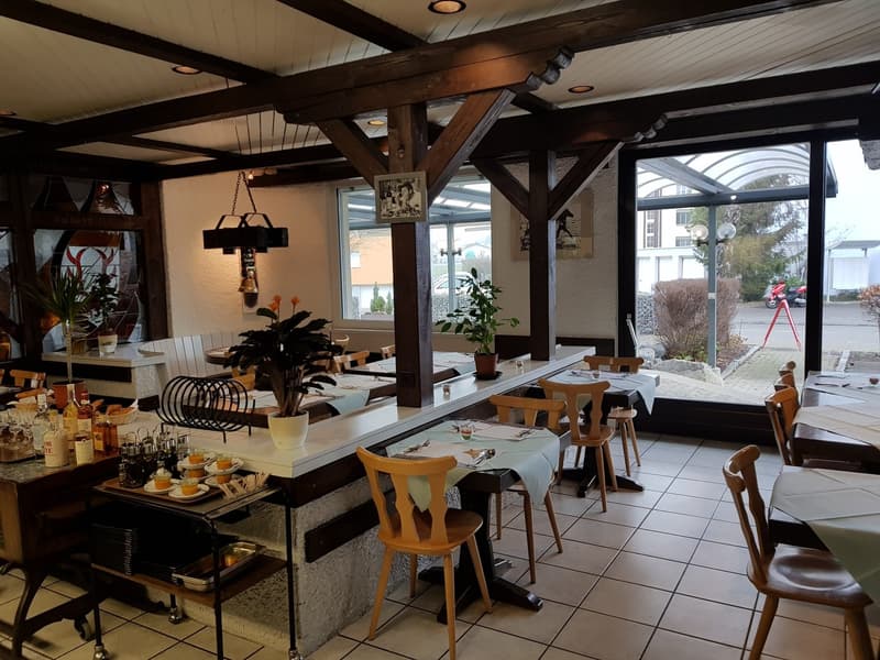 Modern indoor restaurant with large windows, wooden furniture, tiled flooring, various tables and chairs, decorative plants, and a bar counter with bottles and condiments.