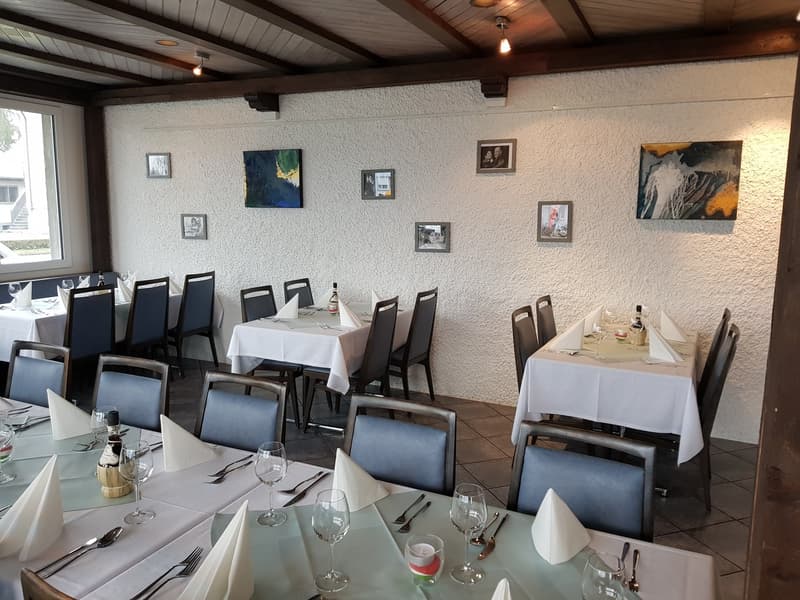 Interior of a restaurant with round tables set for dining, wooden tables, blue chairs, pictures on the wall, tiled floor, lights mounted on the ceiling.