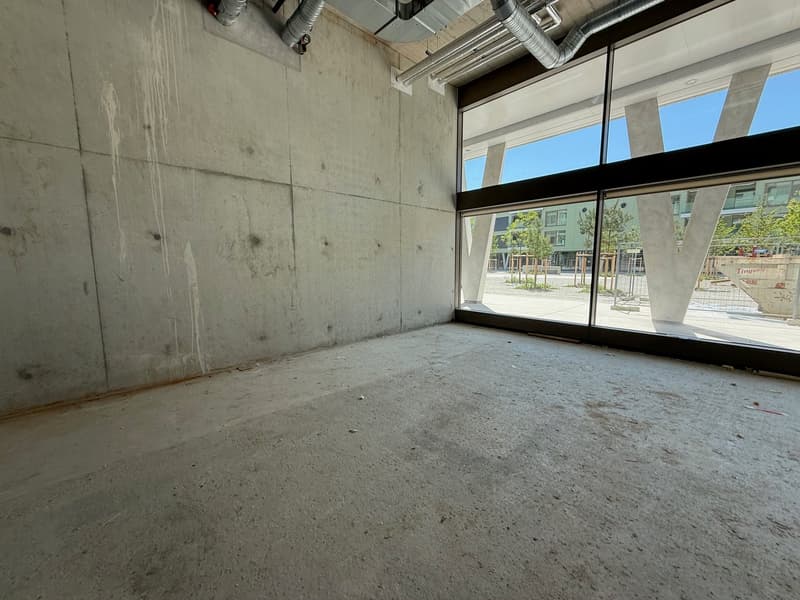 The image shows a large, open commercial space with concrete walls and floors. The space has large windows that provide a view of the outdoor area. The space appears to be in a state of construction or renovation, with exposed ductwork and unfinished surf