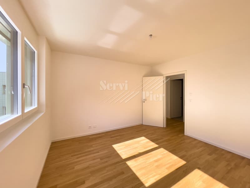 Empty room with hardwood flooring, white walls, and a window. The room has a door leading to another area.