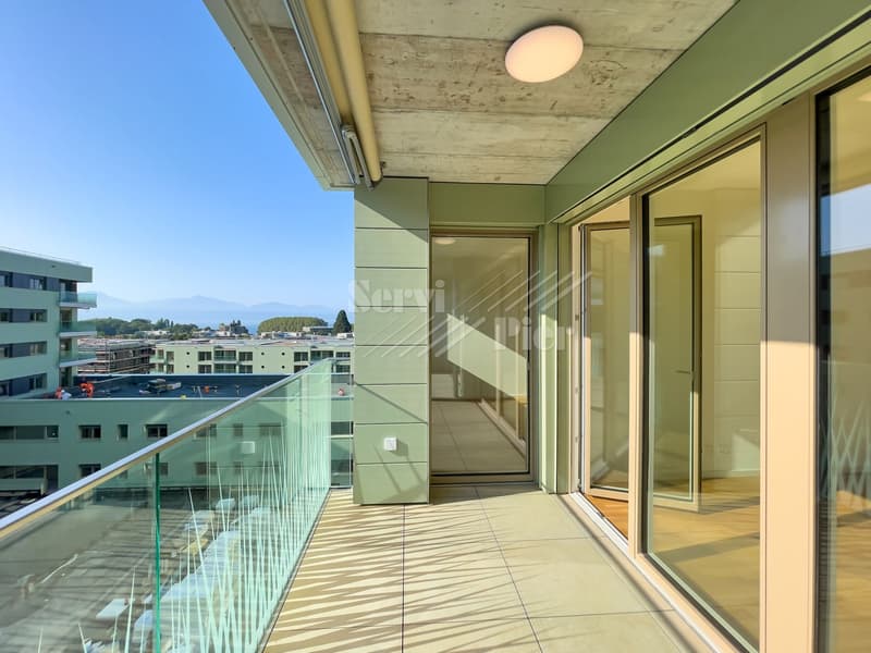 Spacious balcony with glass railing, wooden flooring, sliding glass doors, concrete ceiling, and views of surrounding buildings and landscape.