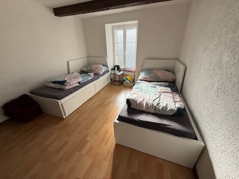 Two beds, wooden floor, white walls, white headboards, small window, night table, rug, storage box
