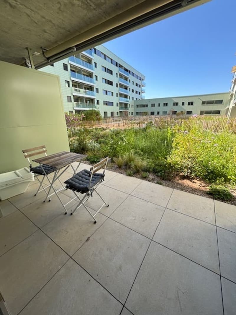 Balcony with table and chairs, tiled floor, green plants, buildings in the background