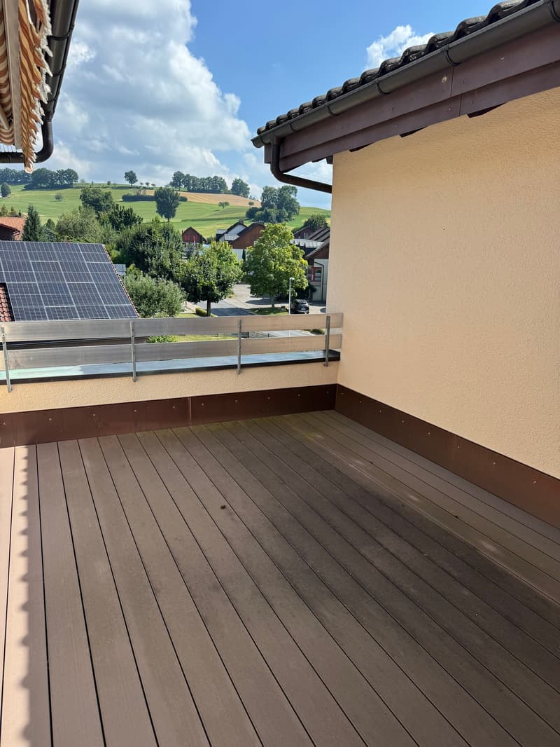 Balcony deck with wooden flooring, metal railings, overlooking a scenic landscape. It has a metal bench seat for comfort. There are solar panels and a tree with lush foliage in the background, along with a picturesque view of hills and houses. The sky is