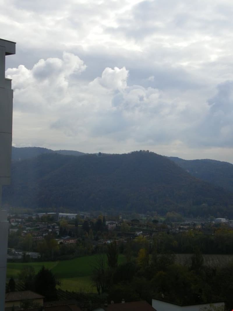The image shows a scenic view of a mountainous landscape with rolling hills and a cloudy sky. The foreground includes some buildings and greenery, while the background features a range of hills and mountains.