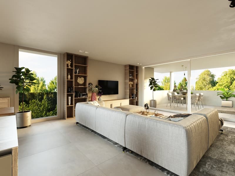 Spacious open-plan living room with large windows overlooking a balcony and lush greenery outside. The room features a comfortable gray sofa, a wall-mounted TV with built-in shelving, and potted plants adding a touch of nature. The dining area is visible