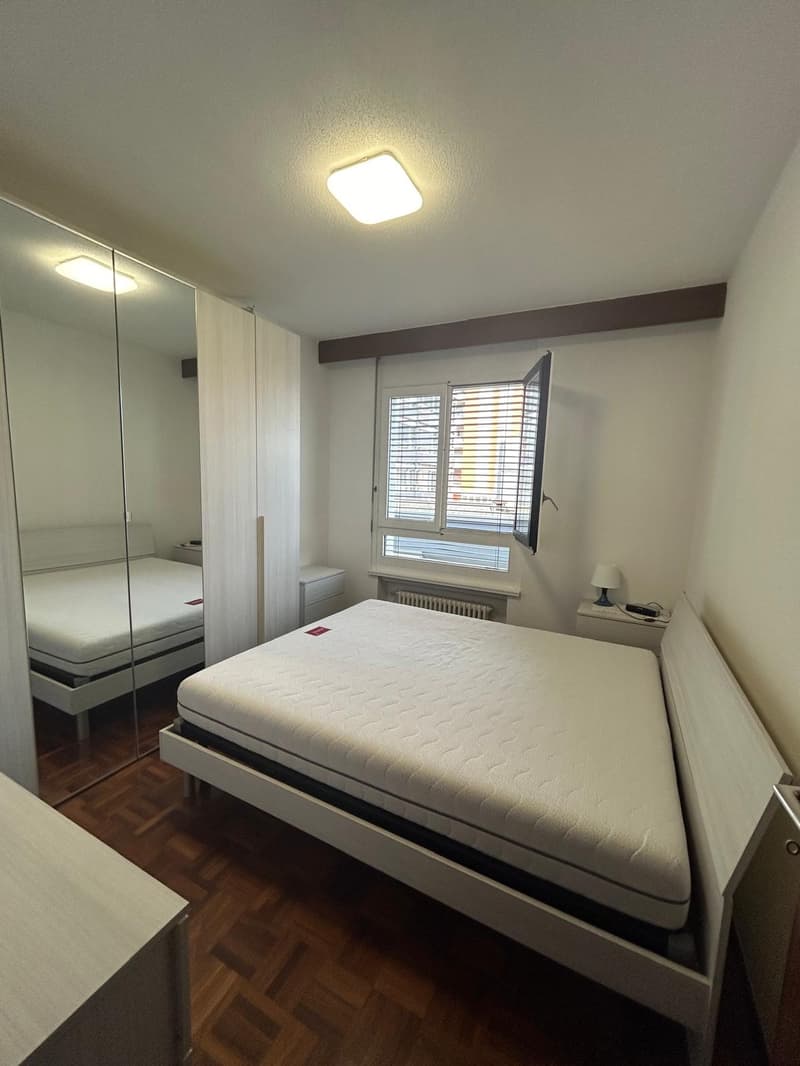 Spacious bedroom with white walls, hardwood floor, full-sized bed, mirrored wardrobe, bedside table, lamp, and window with wooden shutters. Another bedroom with a bed and a nightstand is visible in the mirror.