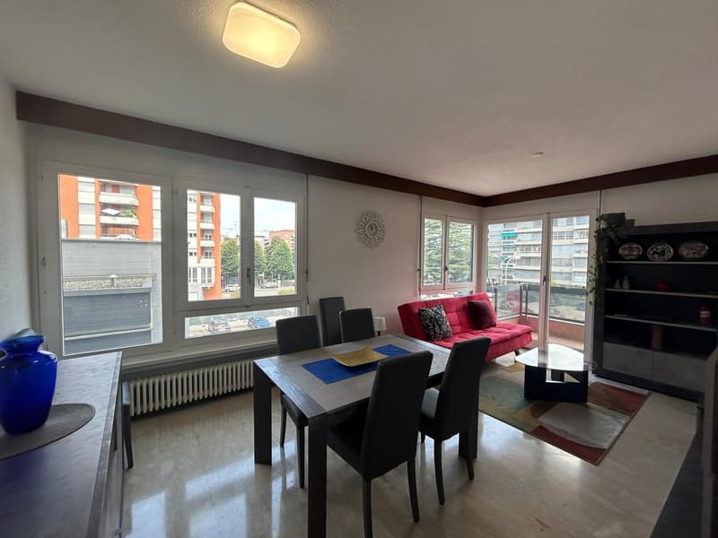 Dining area with a dining table, chairs, couch, clock on the wall, shelves, radiator, windows with balcony, light blue vase, carpet, and a radiator