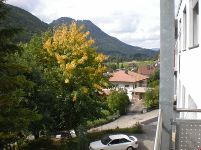 House with red roof, white walls, surrounded by trees and mountains, car parked in front