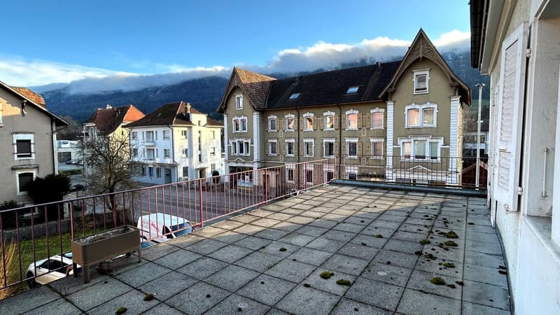 A view of an outdoor balcony or terrace, fenced with metal railings and located on the top floor of a building. The balcony has a tiled floor and overlooks a parking area where several cars are parked. In the distance, there are trees and a scenic mountai