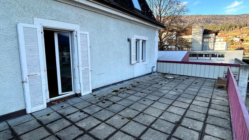 Terrace with white shutters, tiled floor, glass door, balcony railings, outdoor seating, distant buildings and hills