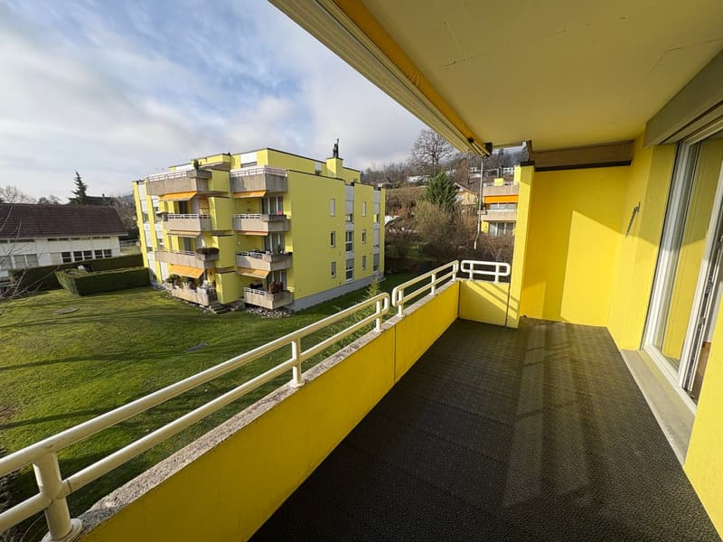 A modern yellow apartment building with balconies and awnings in a residential area, a well-maintained green lawn, and distant trees and buildings