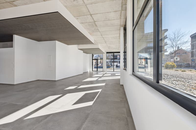 Large open space with concrete flooring, white walls, and large windows providing natural light. The space appears to be a commercial or retail area, with a modern and minimalist design.