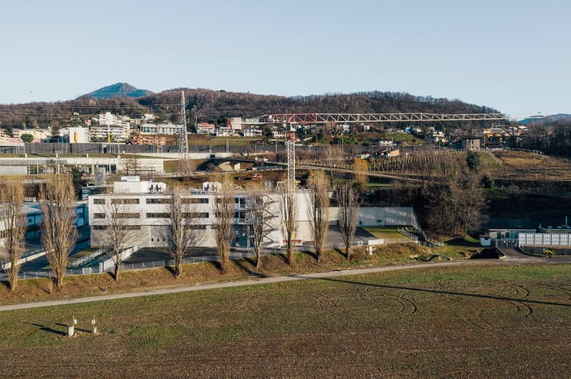 The image shows a cityscape with a mix of residential and commercial buildings, a large bridge or overpass structure, and a hilly, forested area in the background. There appears to be a parking area or lot in the foreground.