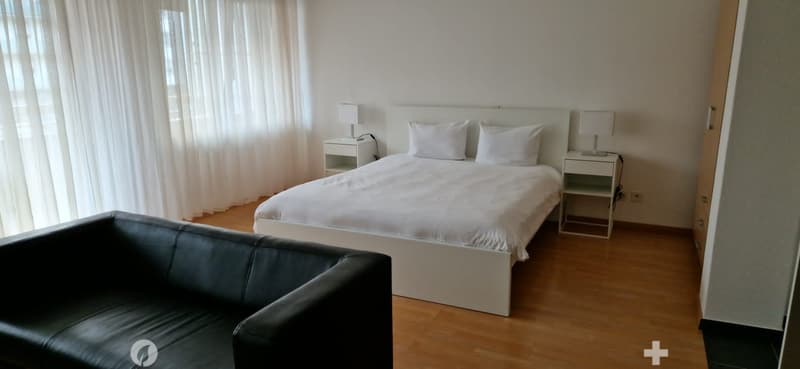 A bedroom with white bed, two white nightstands with lamps, white pillows, black leather couch, and wooden floor.