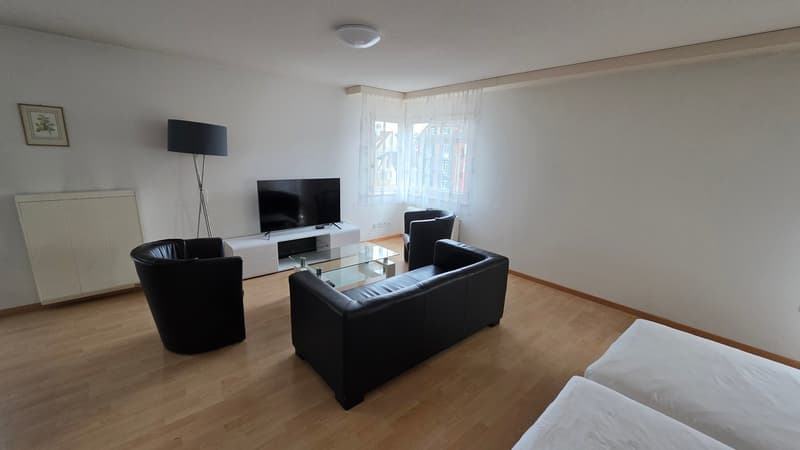 white walls, brown wood flooring, black leather couch, glass table, white tv stand, black TV, three black chairs, large window with white curtains