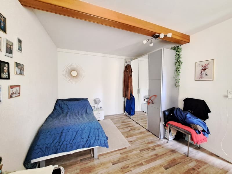 Bedroom with hardwood flooring, white walls, wooden beams on the ceiling, a large bed with a blue comforter, a wardrobe, and some decor items like framed artwork and a plant.
