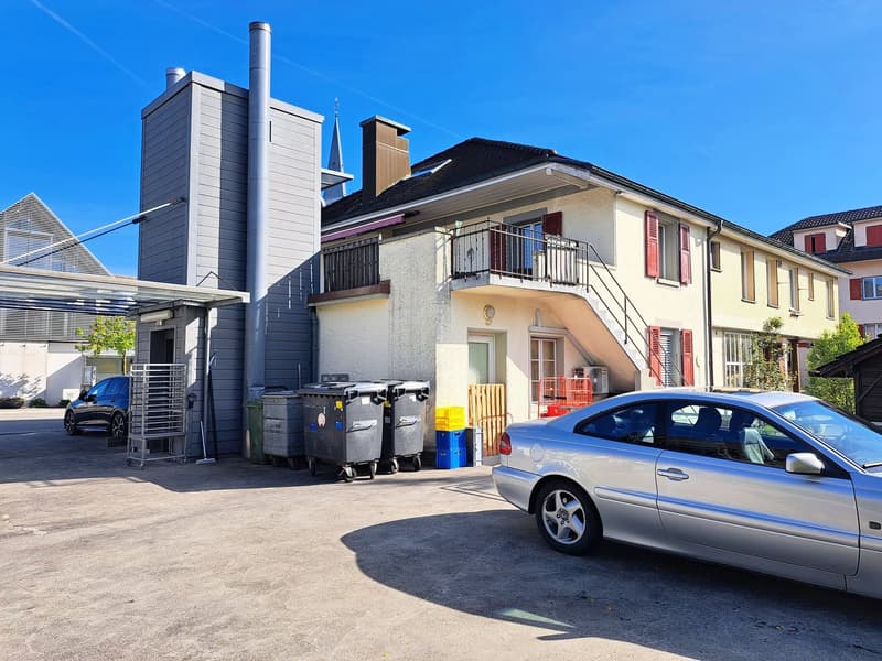 2-story building with a modern, gray exterior and a tall chimney. The building has balconies and there is a parked car in the foreground.