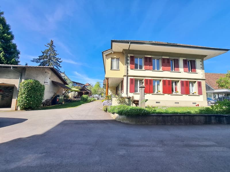 Two-story house with yellow exterior, red shutters, and a balcony. There is a paved driveway and landscaped garden in the front of the house.