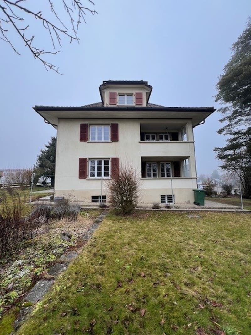 A two-story house with white walls and red shutters, a tiled roof, a balcony, a garden with green grass, and a green trash bin on the left side of the house.