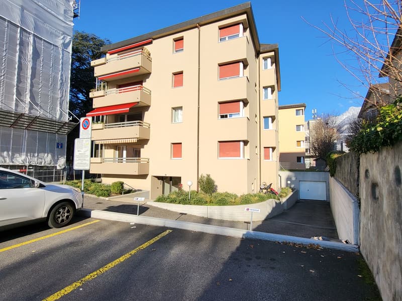 A residential apartment building, 4 stories, beige paint, multiple balconies, underground garage, plants in front, car parked in front