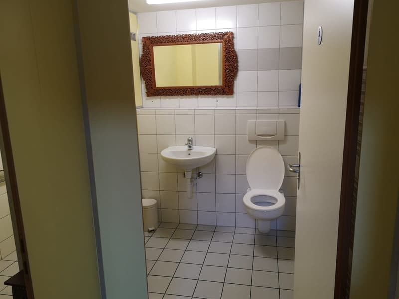 White tiled floor and walls, white sink, toilet, wooden mirror with decorative frame, white toilet paper holder, small waste bin, door opening.