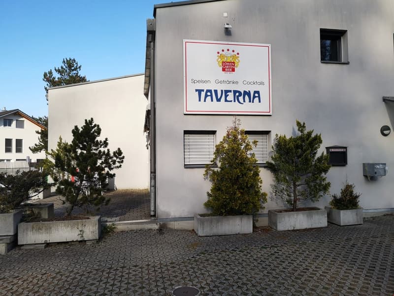 white exterior, grey paving in front, rectangular sign with Taverna logo, window with blinds, shrubs in planters