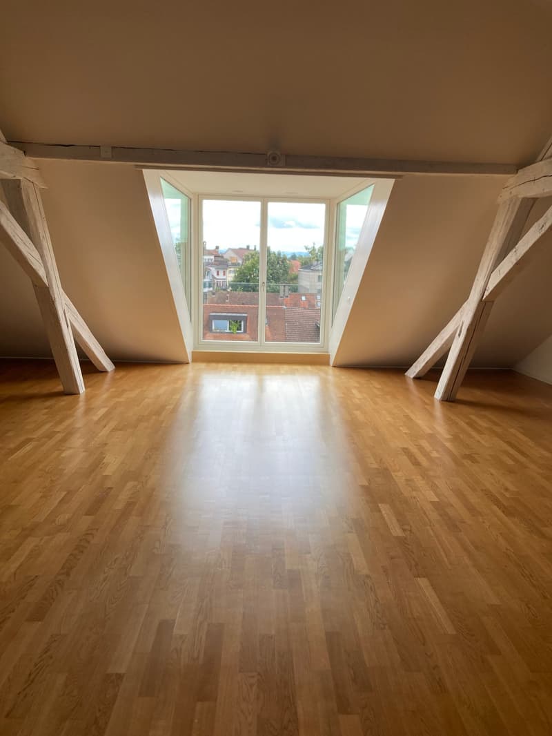 Spacious attic room with hardwood flooring, large windows providing natural light and a view of the surrounding buildings.