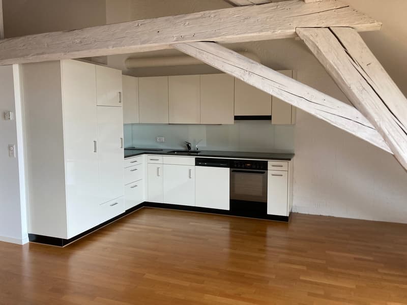 The image shows a modern kitchen with white cabinets, a black countertop, and a built-in oven and stovetop. The kitchen has a wooden beam running across the ceiling, adding a rustic touch to the space. The floor is made of hardwood planks.