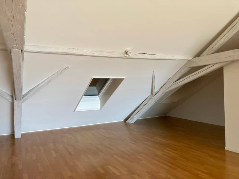 Spacious attic room with wooden floors, white walls, and slanted ceilings with exposed beams. There is a small window providing natural light.