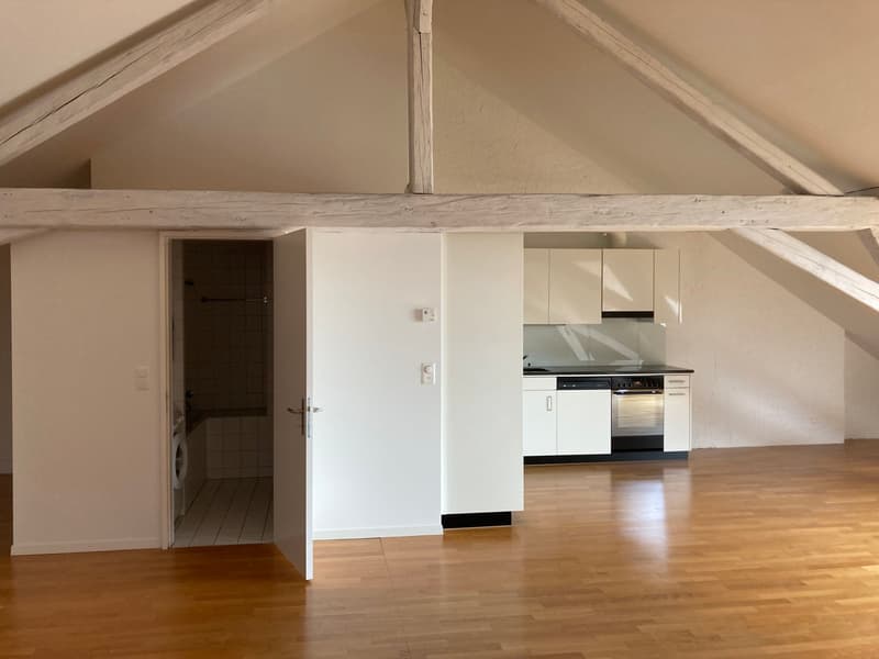 The image shows an open-plan studio apartment with a vaulted ceiling and exposed wooden beams. The apartment has a kitchen area with white cabinets and appliances, and a hardwood floor throughout. There is a door leading to what appears to be a bathroom o