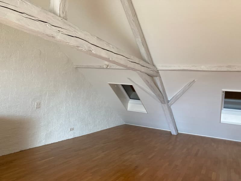 Attic room with exposed wooden beams, white walls, and hardwood flooring. The room has a small window and appears to be a storage or living space.