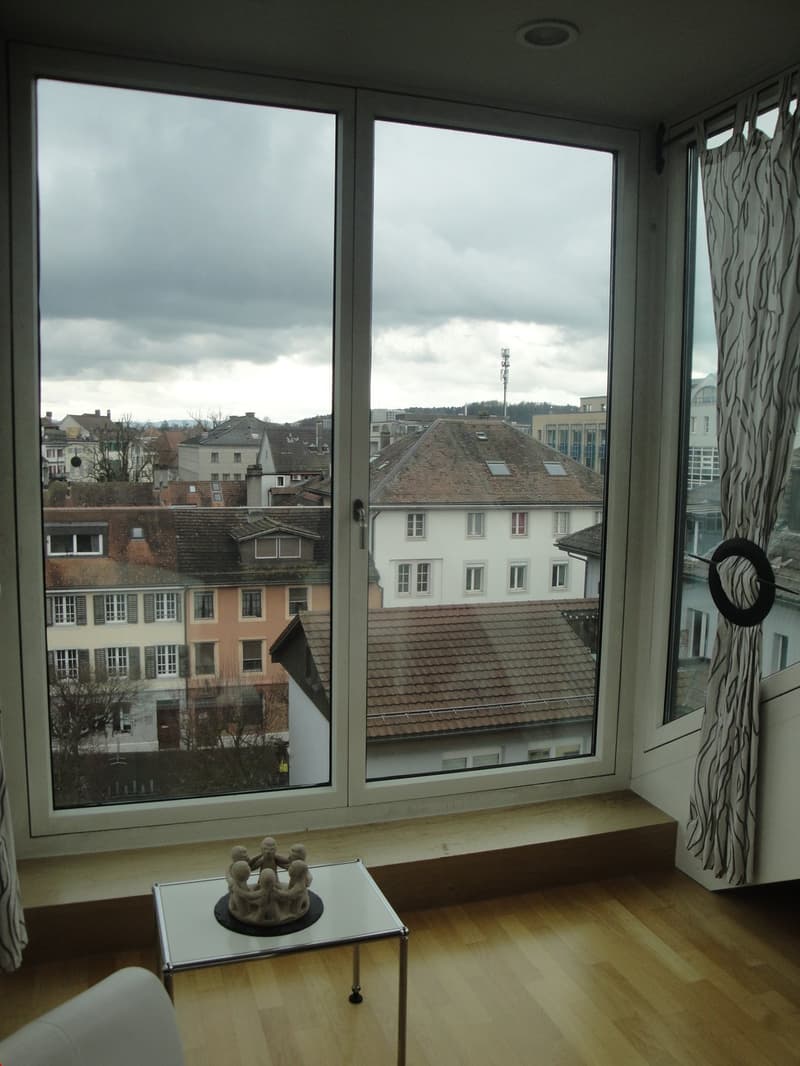Large windows with a view of the city skyline, wooden floor, curtains, and a small table with a decorative sculpture on it.