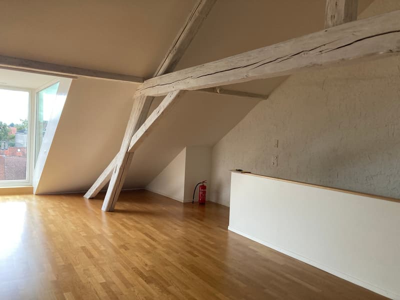 Spacious attic room with wooden beams, hardwood flooring, and white walls. The room has a large window providing natural light. There is a built-in cabinet or storage unit along one wall.