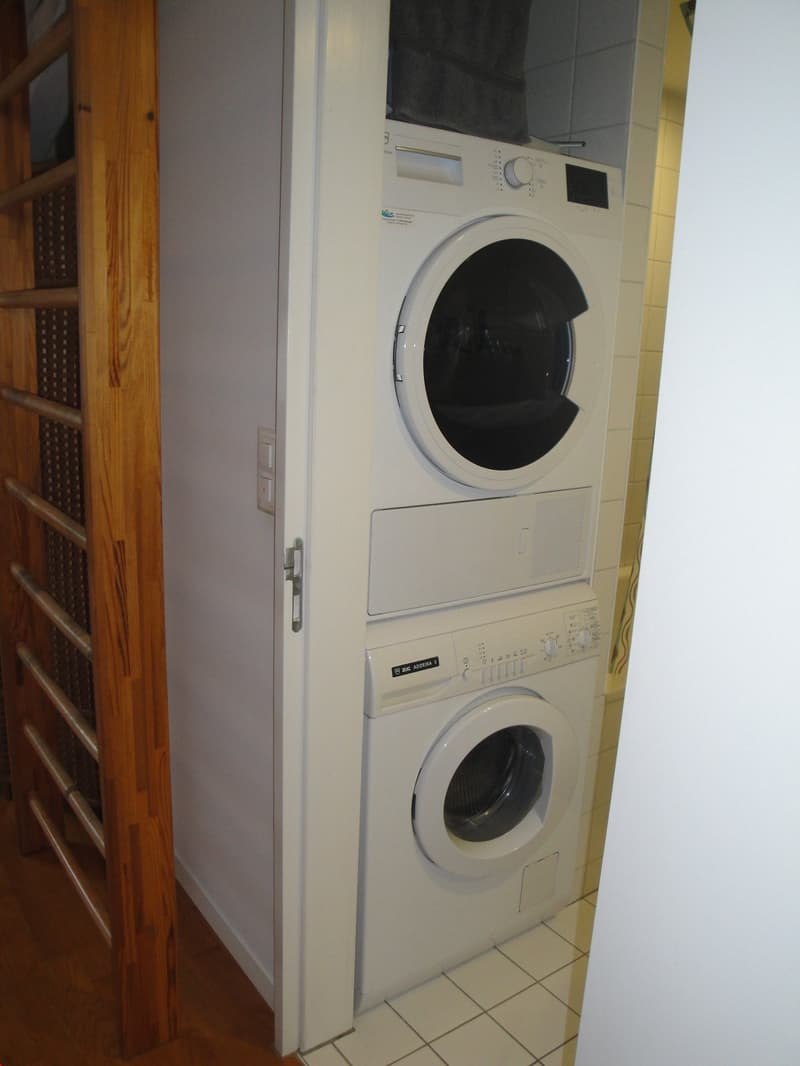 The image shows a stacked washer and dryer unit in a tiled bathroom or laundry room. The washer and dryer appear to be white appliances, and there is a wooden shelving unit next to them.