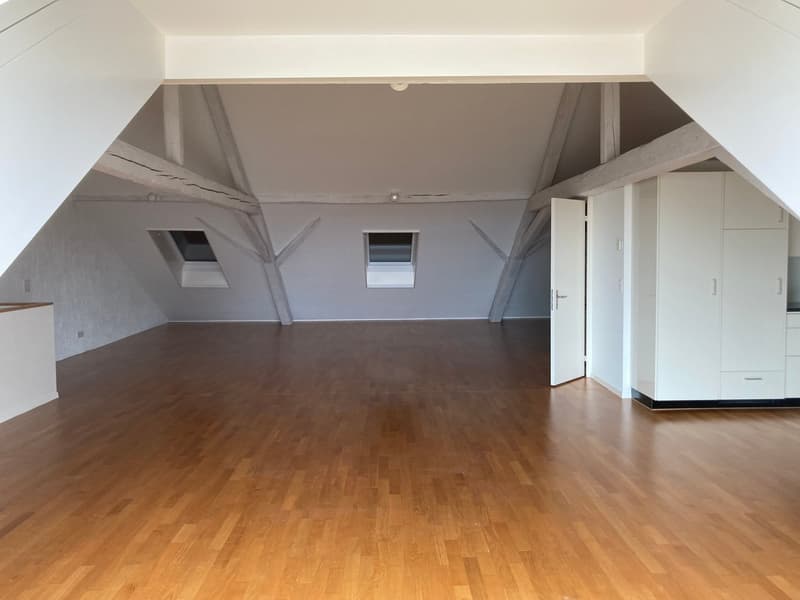 Large open space with wooden flooring, white walls, and slanted ceilings with exposed beams. The room has two small windows and a white door leading to another area.