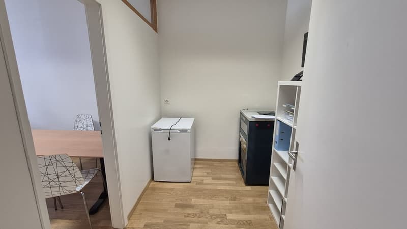 small room with wooden floor, white walls, table, chairs, small fridge, cabinet, shelves