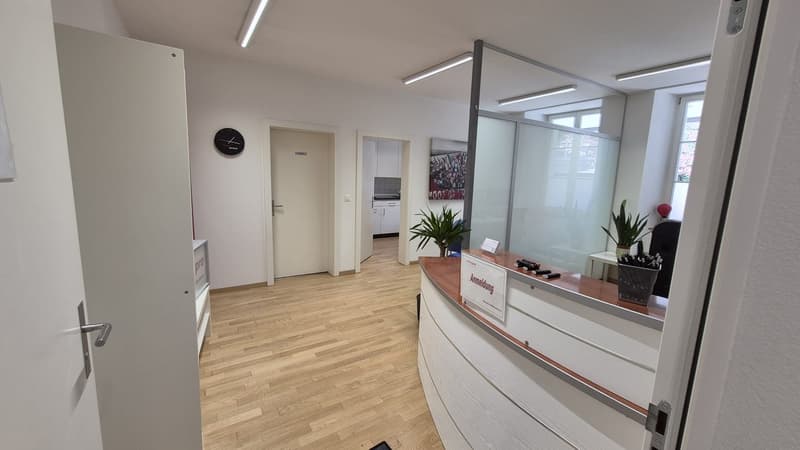 Reception area with wooden floor, white walls, glass partition, plants, and a clock on the wall.