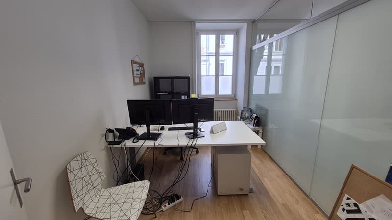 Office room with desk, two monitors, keyboard, mouse, chair, bulletin board, radiator, window, TV