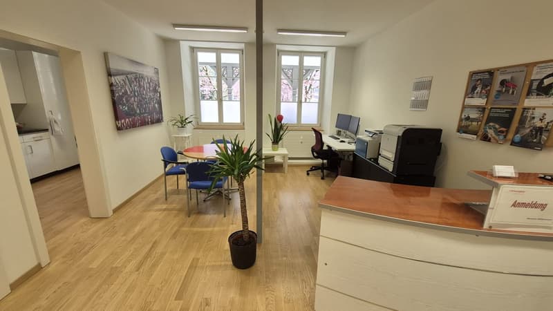 Wooden floor, white walls, round table, blue chairs, white cabinets, desk, computer, printer, potted plants, windows, bulletin board
