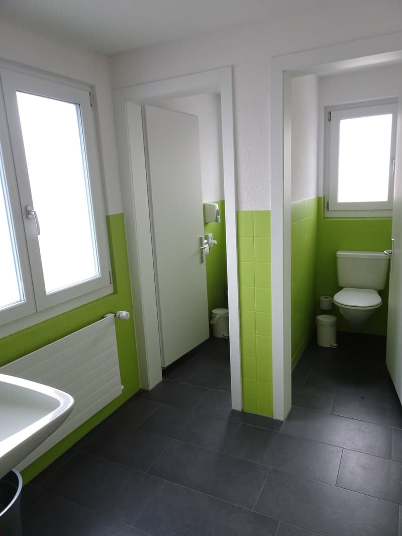 Spacious bathroom with white walls, green tile accents, black tile flooring, toilet, sink, and a washing machine