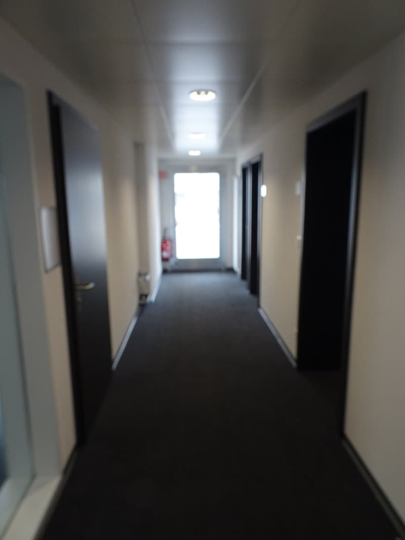 The image shows a long, dimly lit hallway with white walls and a dark carpet floor. There are several doors lining the hallway, suggesting this is a multi-room space. The lighting appears to be provided by recessed ceiling lights, creating a somewhat inst