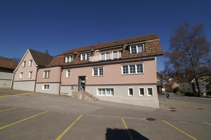 A multi-story building with a brown tiled roof, white windows, and a parking lot in front. The building appears to be a residential or commercial structure located in a town or city setting.