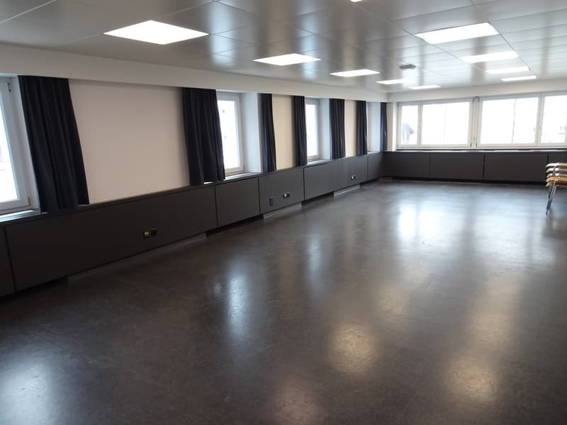 Large open room with concrete flooring, black curtains covering the windows, and recessed lighting in the ceiling. The room appears to be set up for events or meetings, with storage cabinets lining the walls.